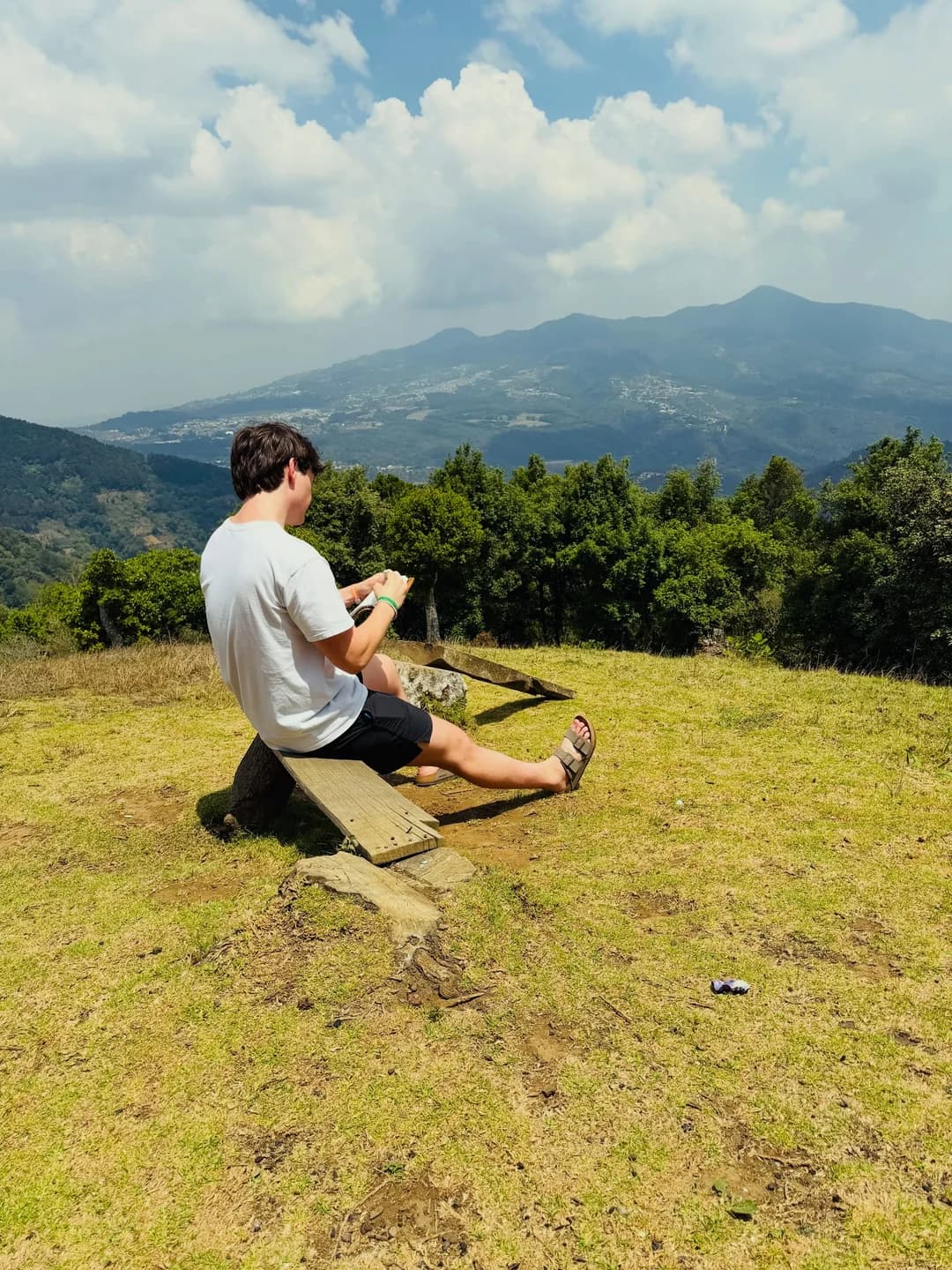 Grant sitting on a wooden bench on a grassy hillside in Guatemala, with mountains behind him.